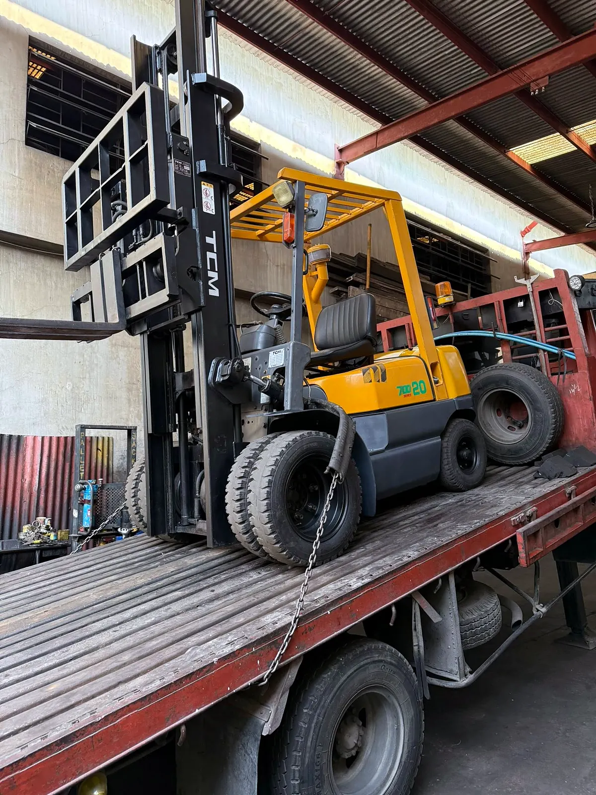 A TCM forklift chained down on a flatbed truck inside a warehouse yard, loaded for delivery