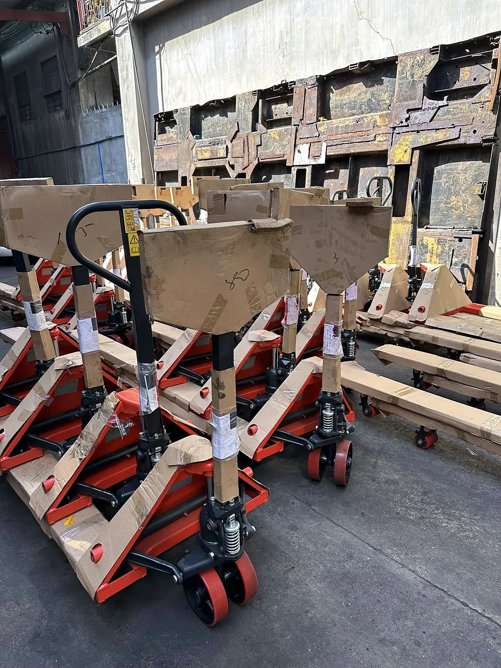 Rows of new hand pallet jacks, still wrapped in cardboard, in a sunlit yard