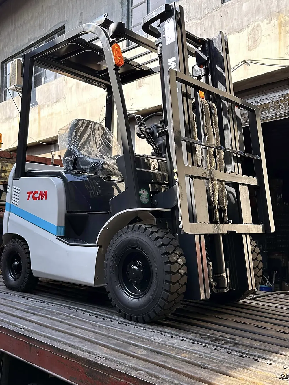 White and blue TCM electric forklift with its seat still wrapped, loaded on a flatbed truck